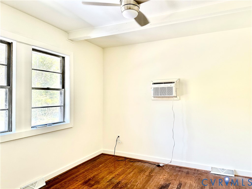Interior room featuring wood-finish flooring, white walls, dual-pane windows, a ceiling fan with integrated lighting, and a wall-mounted air conditioning unit