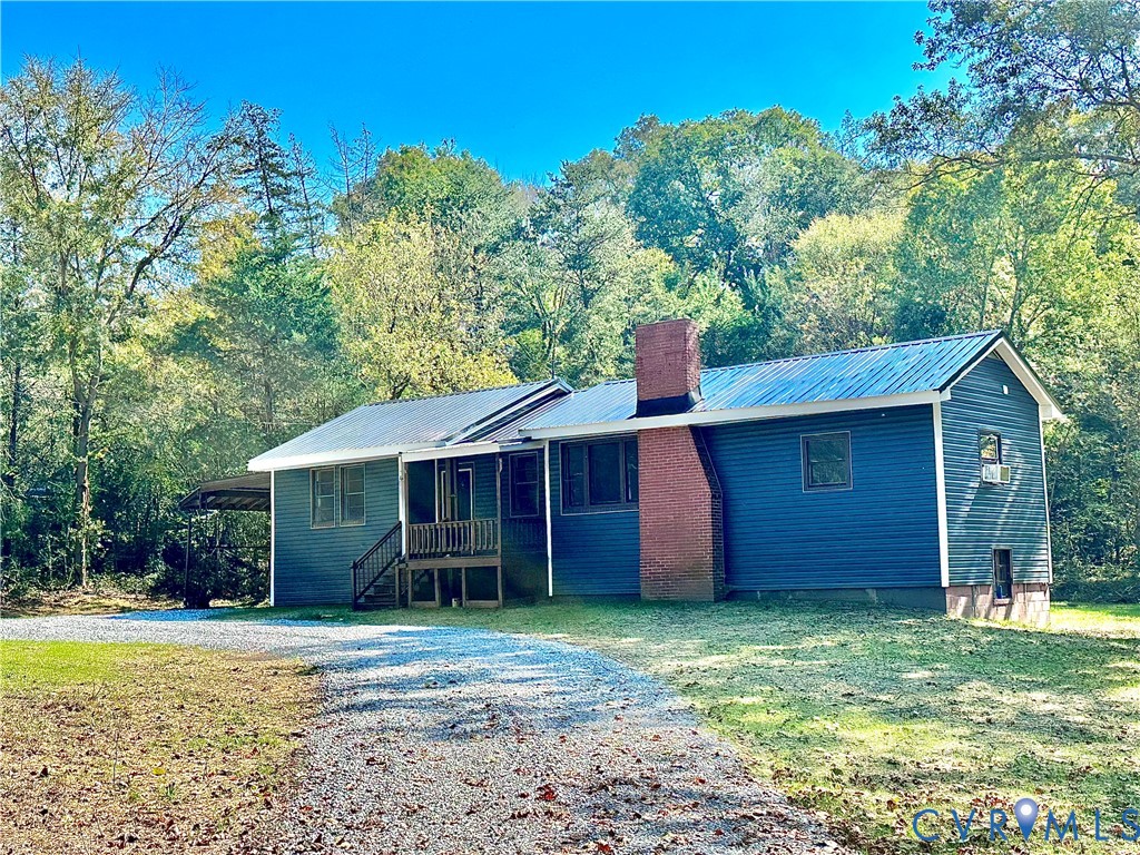 Exterior featuring dark blue horizontal siding, a standing-seam metal roof, a prominent exterior brick chimney, a covered porch, and a gravel driveway