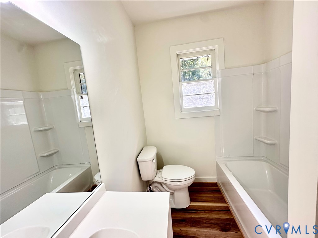 Bathroom featuring wood-finish flooring, a white vanity with a single basin, a large wall-mounted mirror, a tub-shower combination with integrated shelving, and a double-hung window