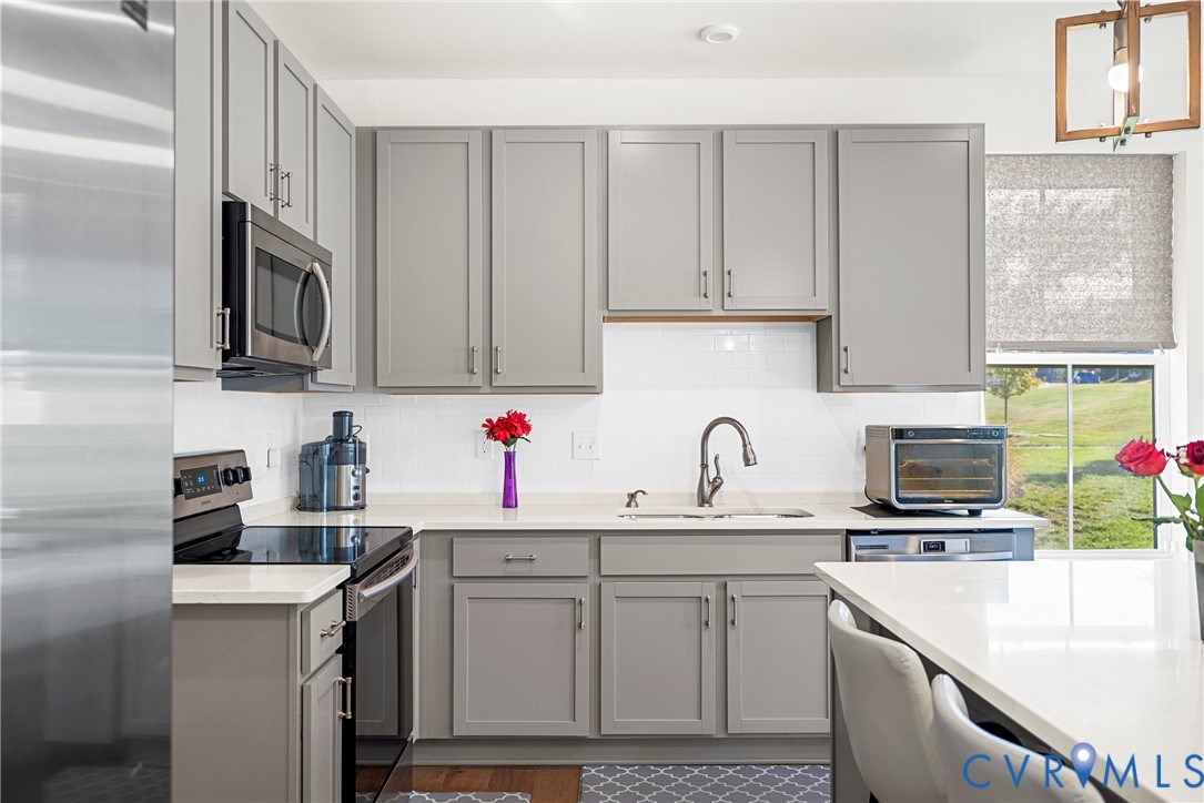 Kitchen featuring grey shaker-style cabinetry, white solid-surface countertops, subway tile backsplash, stainless steel appliances, and a rectangular pendant light fixture
