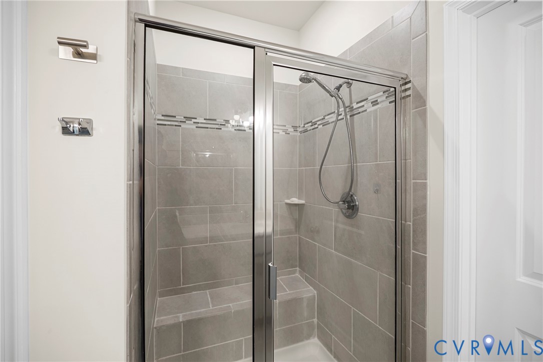 Walk-in shower featuring large-format gray tile surround, decorative mosaic glass tile inlay, integrated corner bench seat, polished chrome hand-held shower fixture, and a framed glass door