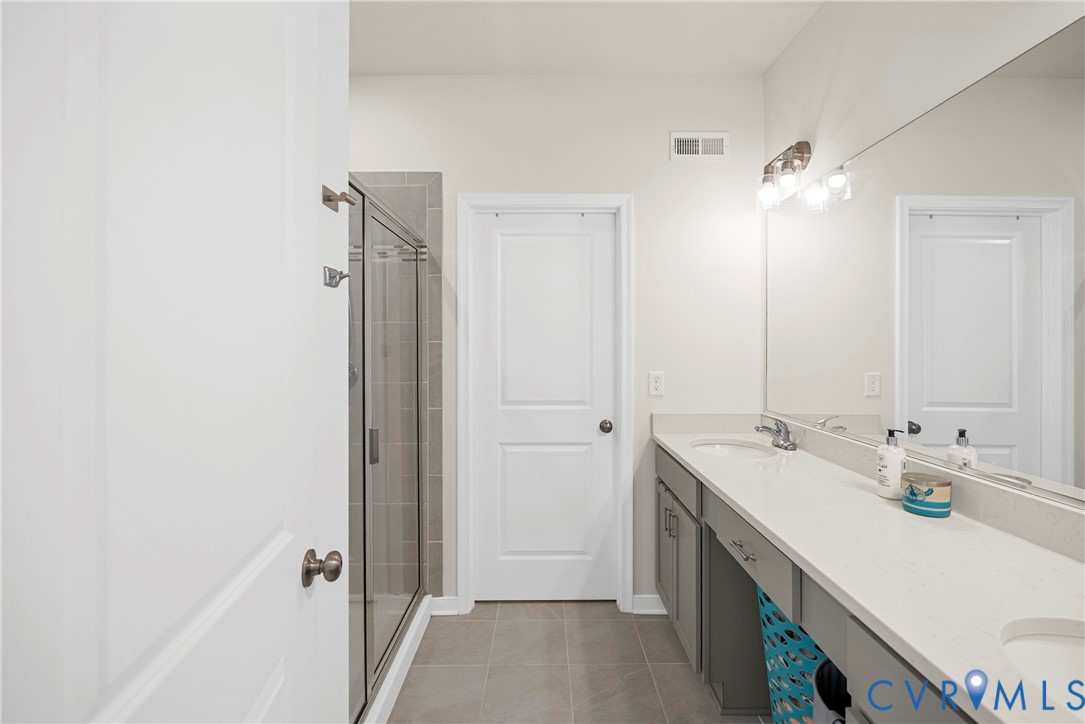 Full-width vanity with a light-toned solid surface countertop and dual undermount sinks, featuring grey cabinetry, a frameless glass-enclosed shower with tile surround, and a wall-to-wall mirror