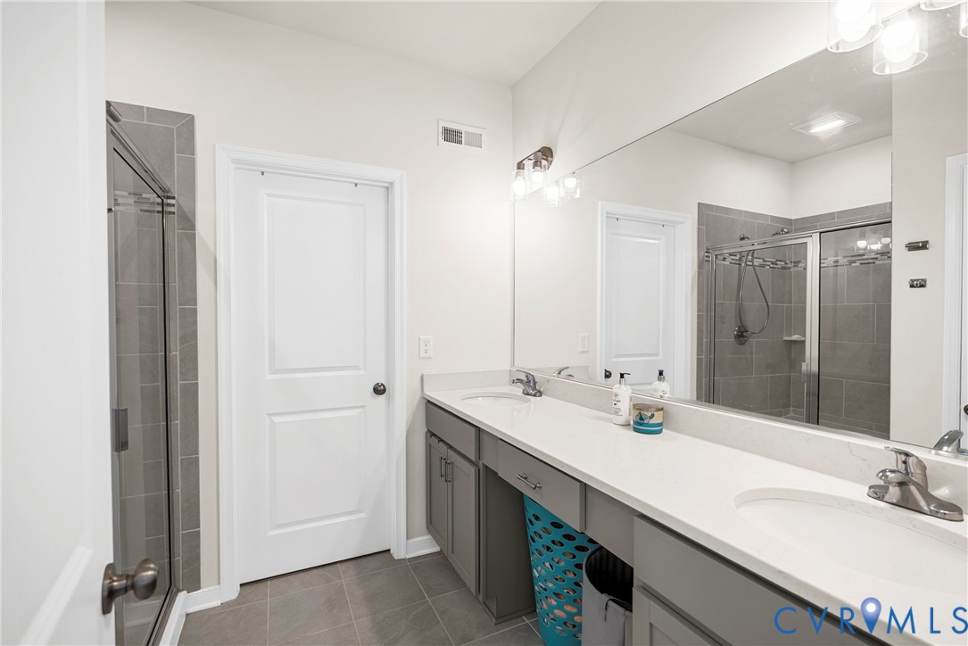 Bathroom featuring a dual-sink vanity with light-toned countertops, gray cabinetry, a large wall-mounted mirror, and a glass-enclosed shower with tile surround