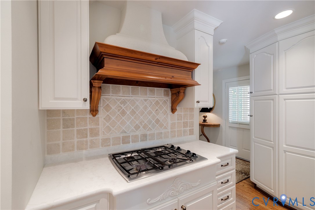 The cooktop area features a stone tile backsplash and lighted wood decorative hood.  Passageway leads to the 2nd entrance area and back hallway.