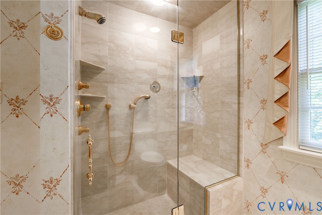 Walk-in shower features floor-to-ceiling tile, brass-finish fixtures, a handheld showerhead, built-in bench seating, and a glass door enclosure.