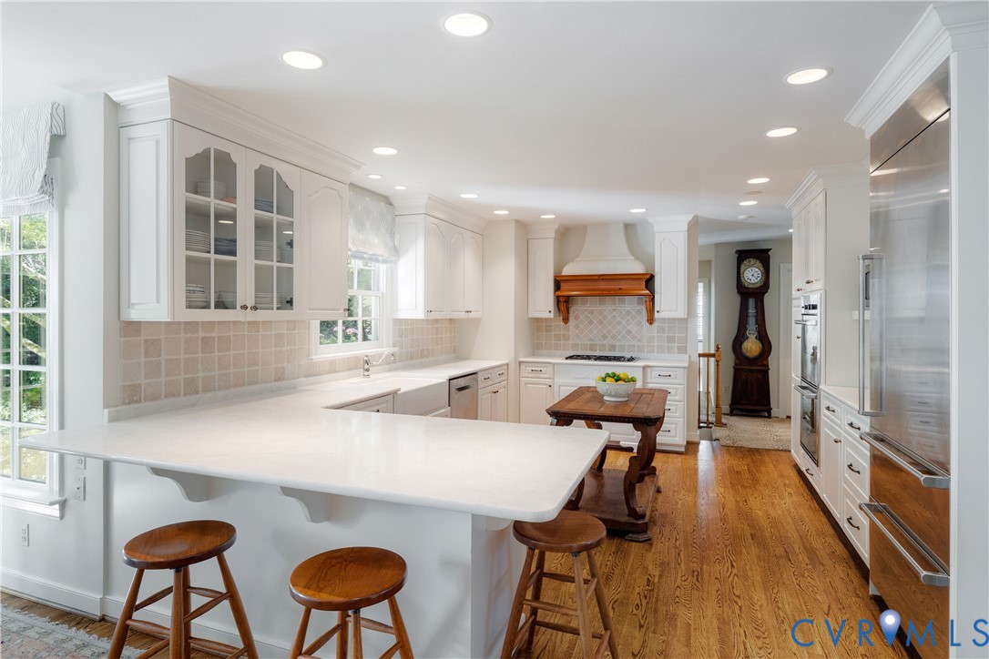 The kitchen has white cabinetry with glass-panel accents, solid-surface counters, a farmhouse-style apron sink, recessed lighting, and gleaming hardwood floors.