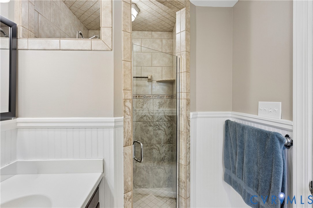 Walk-in shower enclosure featuring tiled surround and stone-finish accent, integrated corner shelving, white beadboard wainscoting, and white vanity countertop