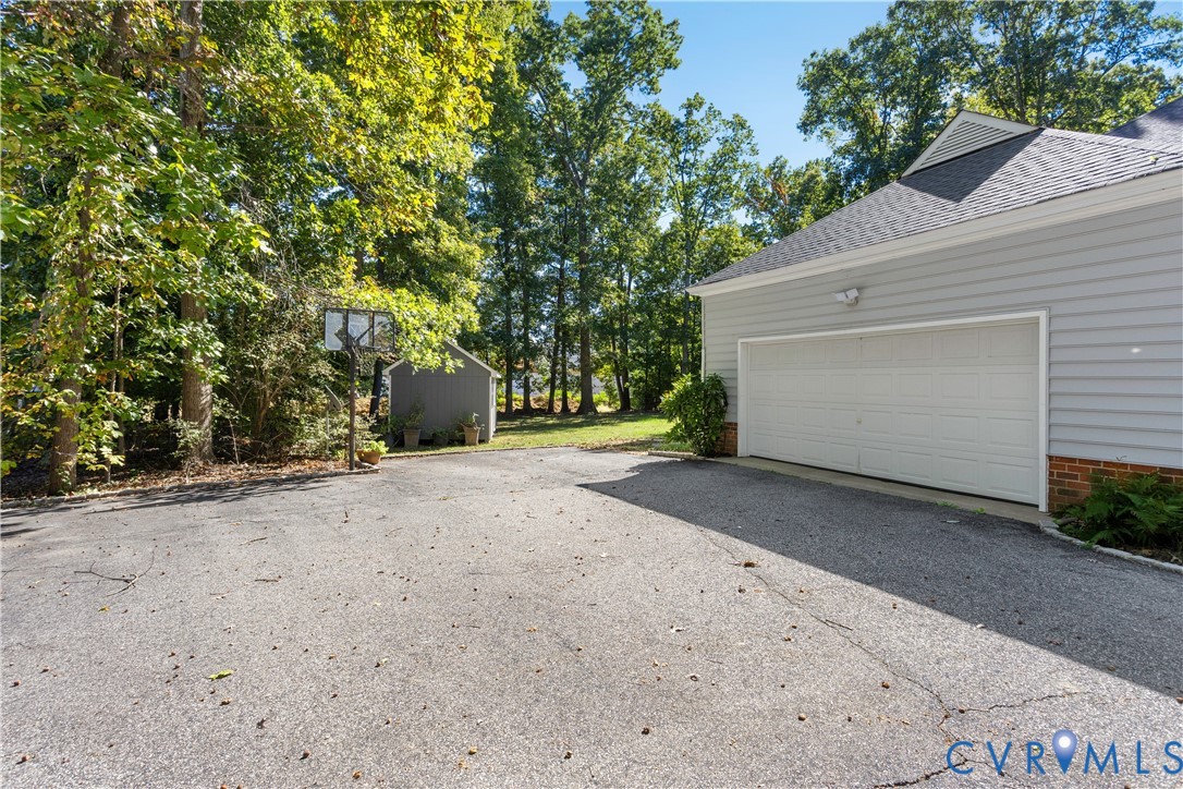 Asphalt driveway featuring a multi-car garage, brick foundation detailing, attached basketball hoop, and a detached storage shed