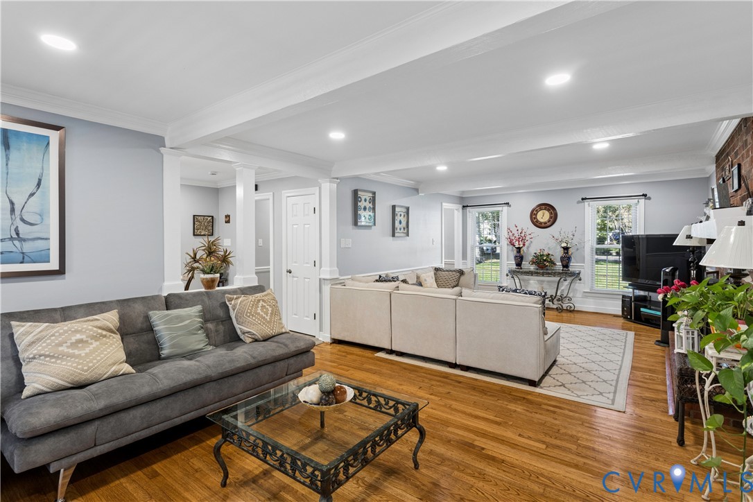 Spacious living area featuring wood-finish flooring, recessed lighting, crown molding, decorative columns, and exposed brick masonry
