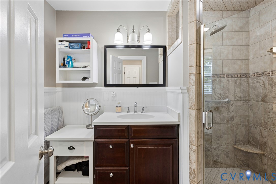 Bathroom featuring a dark wood vanity with a light-colored countertop, three-light fixture above a framed mirror, built-in white wall shelving, and a tiled shower enclosure with a glass door and integrated seat