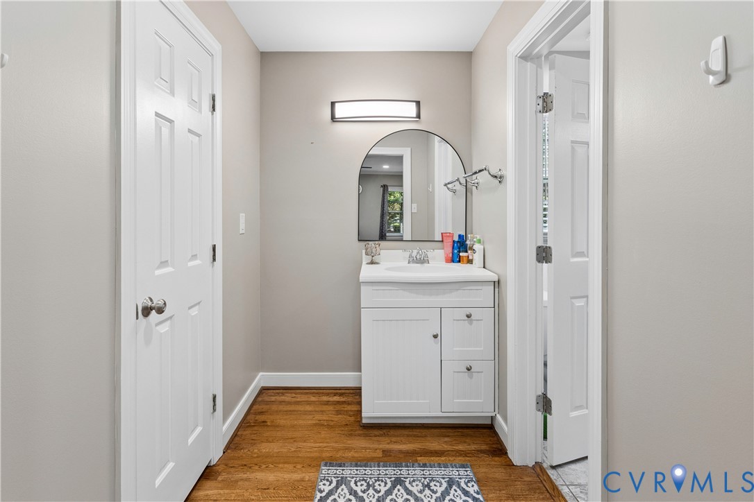 White vanity cabinet featuring an integrated sink, polished chrome hardware, arch-top wall mirror, wall-mounted light fixture, and wood-finish flooring