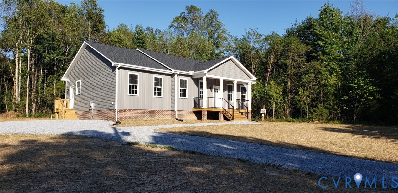 Single-story residence featuring gray vinyl siding, a brick water table, dark shingled roof, a front porch with support columns, and a gravel driveway