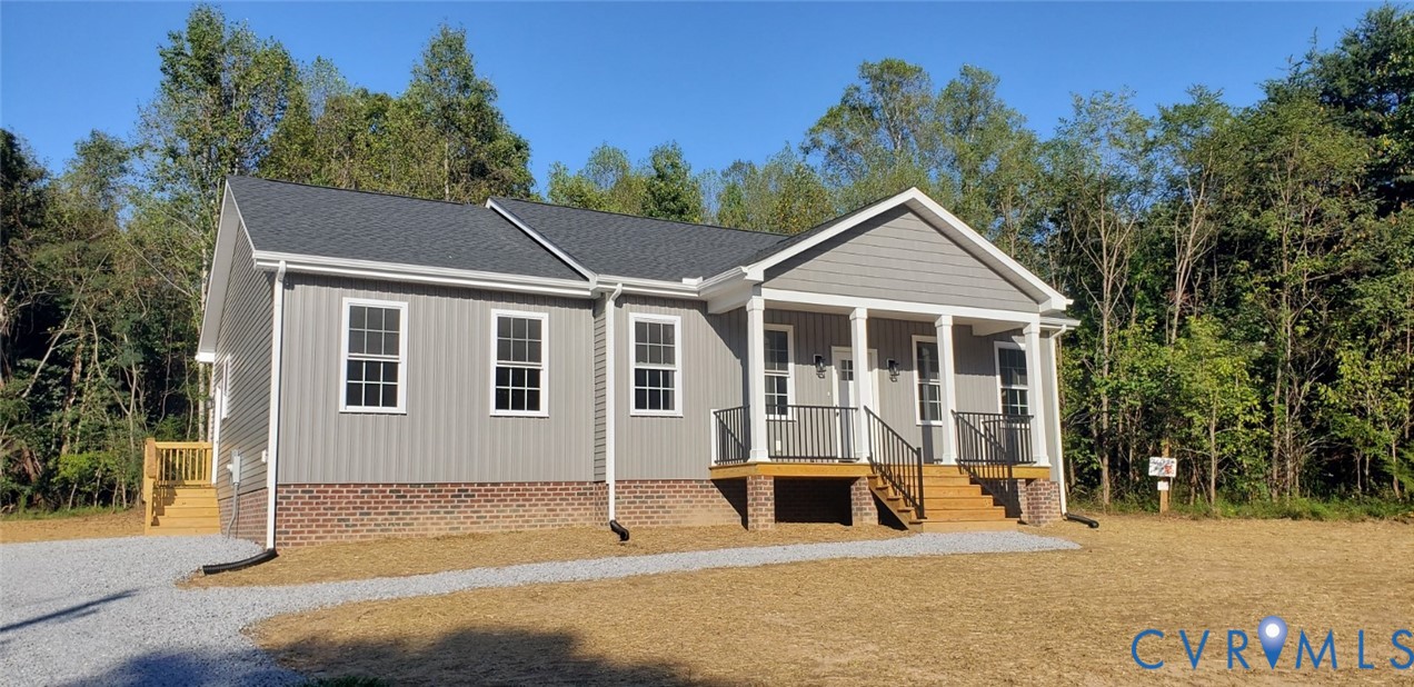 Single-story residence featuring gray vertical siding, a brick foundation, a covered front porch with columns, a metal railing, and a gravel driveway