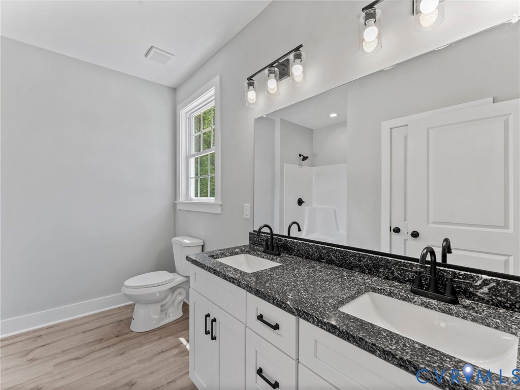 Dual-sink vanity with dark stone-finish countertop, white cabinetry with matte black hardware, wood-finish flooring, and wall-mounted light fixtures