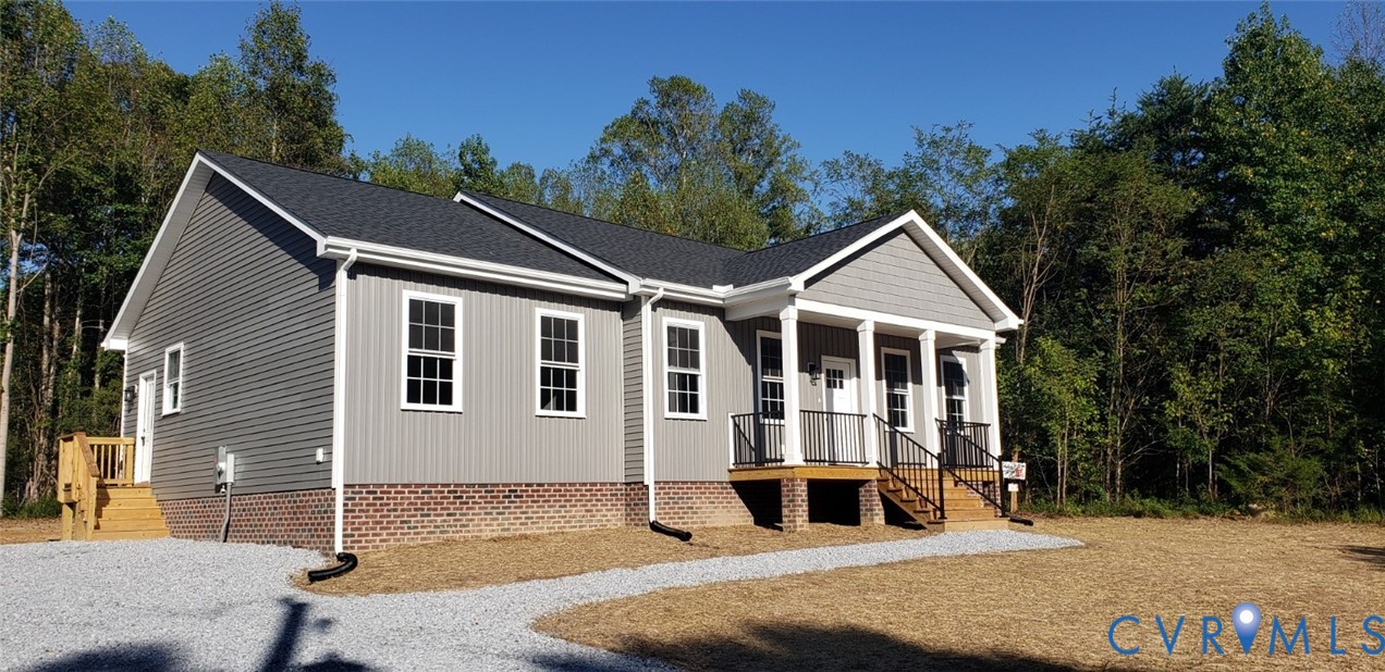 Single-story residence featuring gray vinyl siding, a brick foundation, a covered front porch with white columns, black metal railings, and a gravel driveway