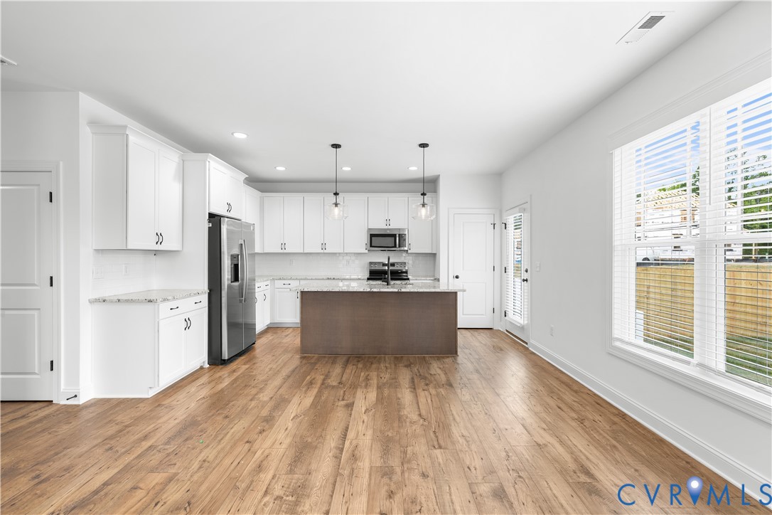 Open-concept kitchen and living area featuring wood-finish flooring, white cabinetry with dark hardware, a center island with a built-in sink, stainless steel appliances, and abundant natural light from a large window