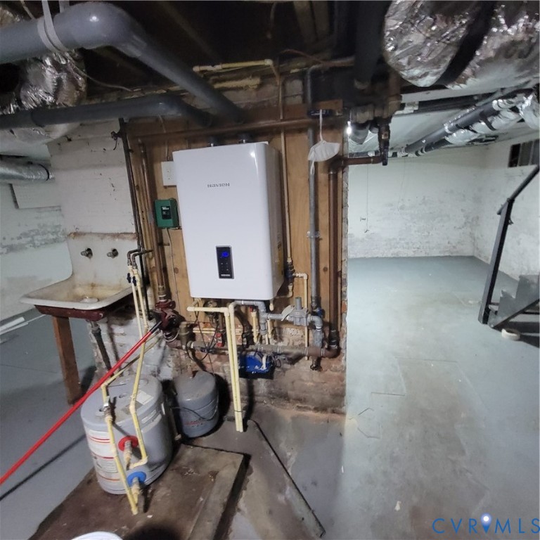 Tankless water heating system featuring a digital display, mounted on a wood backing with exposed utility piping, alongside a wall-mounted utility sink