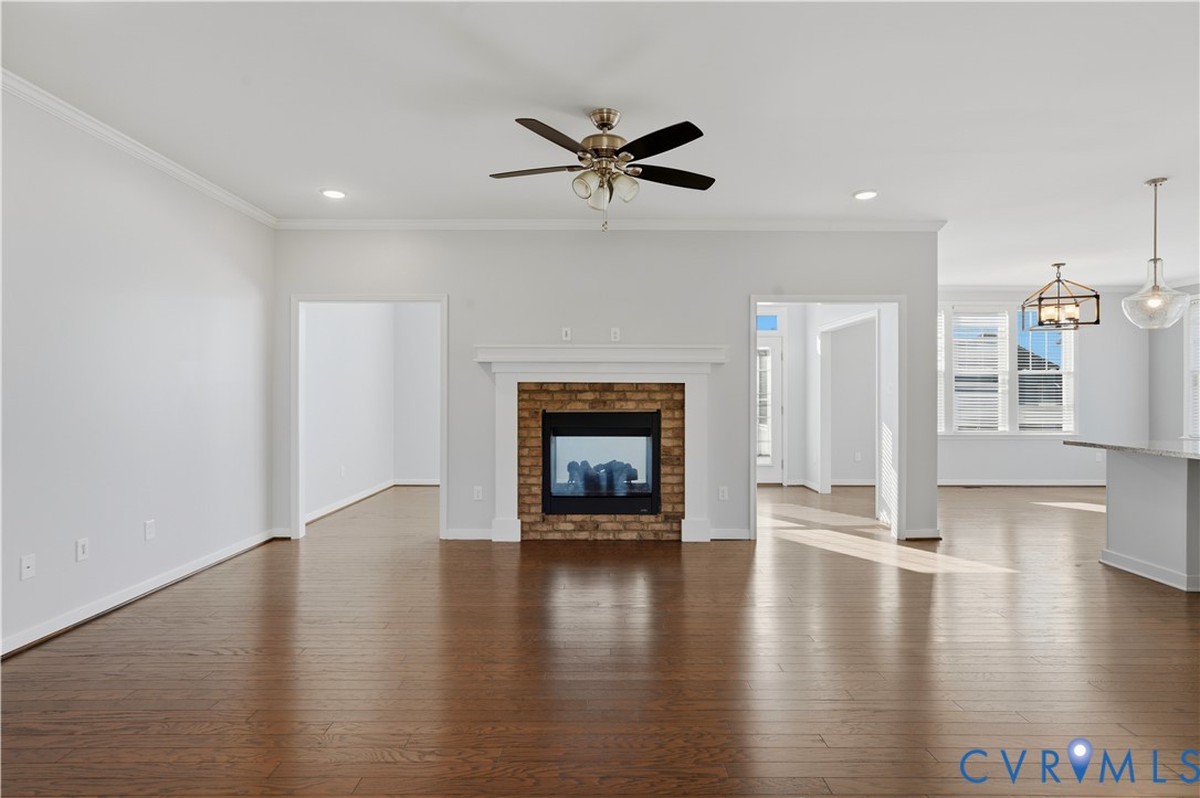 Open-concept interior featuring wood-finish flooring, brick-surround fireplace with a white mantel, ceiling fan with integrated lighting, and modern pendant fixtures