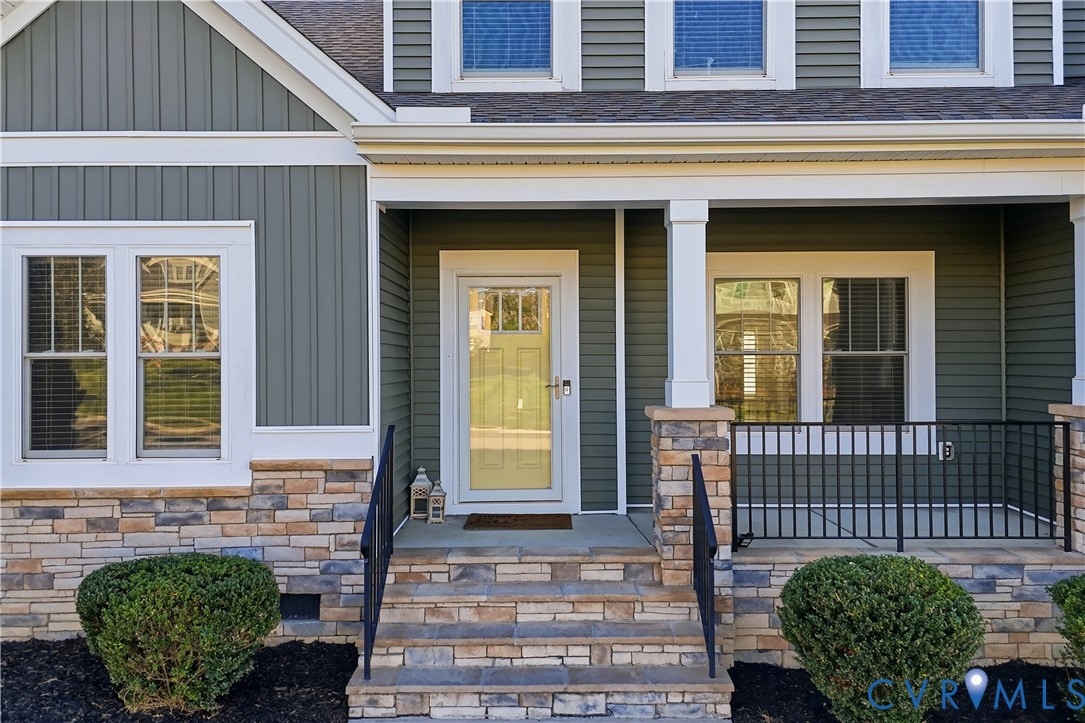 Exterior facade featuring sage green vertical and horizontal siding, stone veneer masonry, covered porch with square columns, and black metal railings