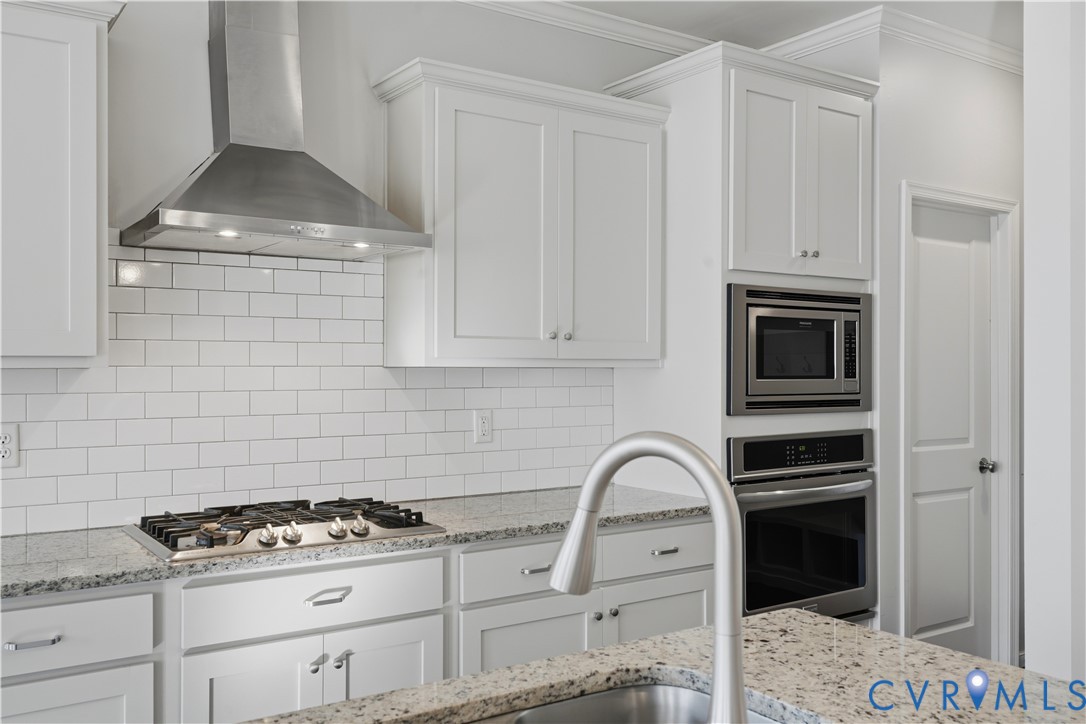 Kitchen featuring white shaker-style cabinetry, stone countertops, a subway tile backsplash, a stainless steel gas cooktop with a matching overhead vent hood, and built-in stainless steel double wall ovens