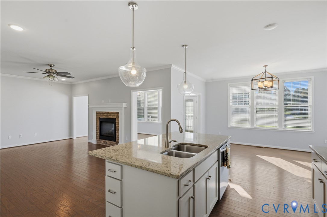 Kitchen island with stone-finish countertop, dual-basin stainless steel sink, wood-finish flooring, fireplace with brick surround, and pendant lighting