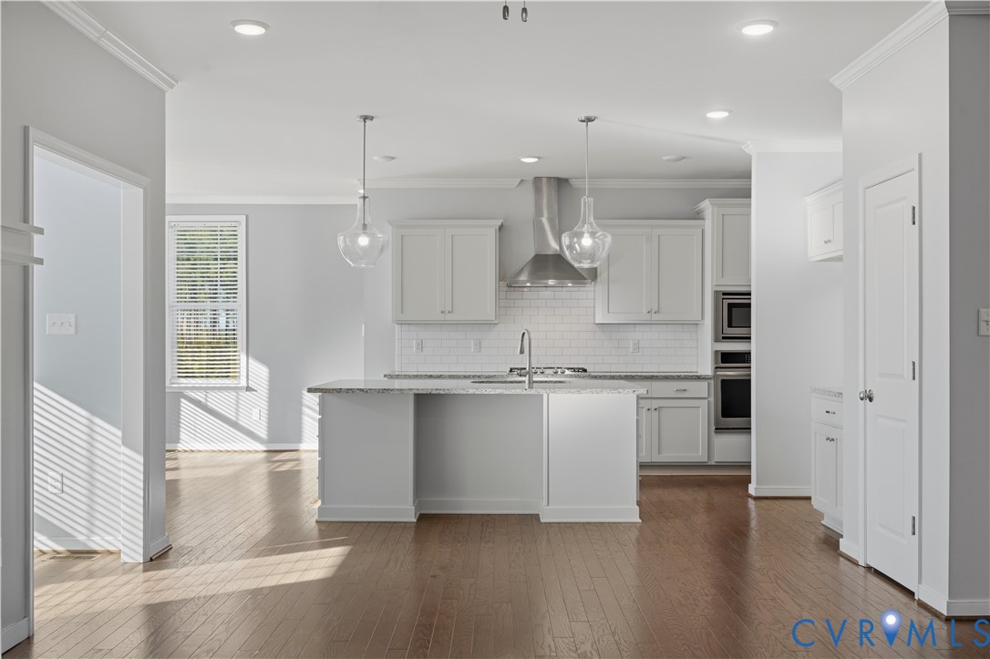 Kitchen featuring an island with a stone countertop, white shaker-style cabinetry, a stainless steel range hood, subway tile backsplash, and wood-finish flooring