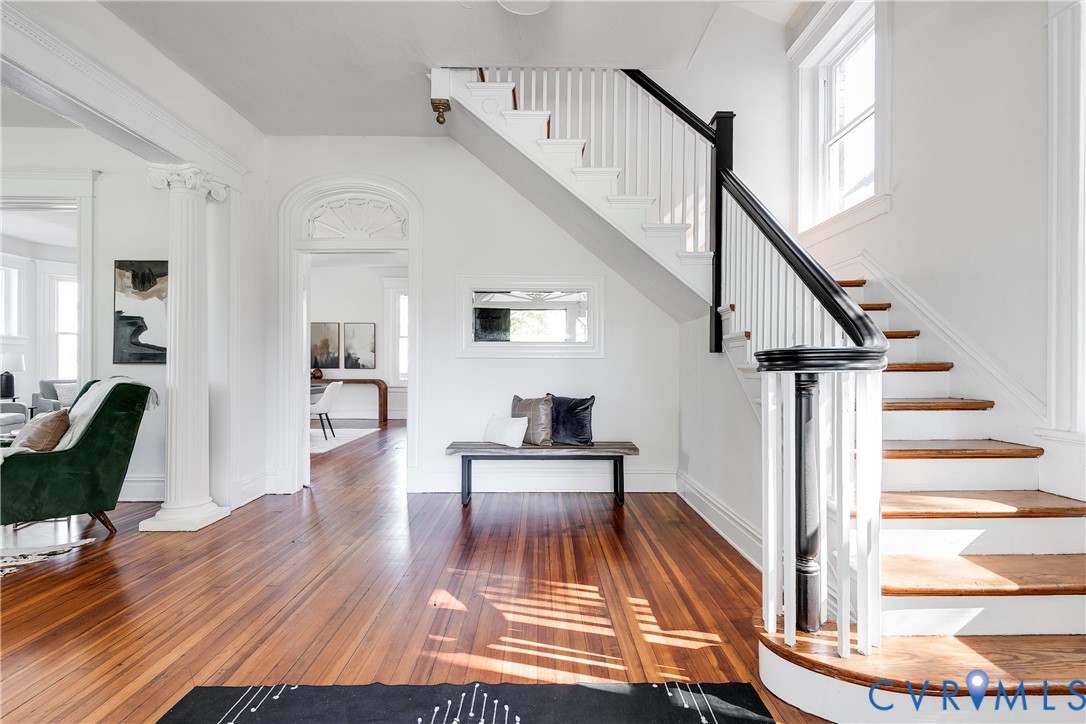 Foyer featuring wood-finish flooring, a classic staircase with a decorative banister, structural columns, and arched doorway detailing
