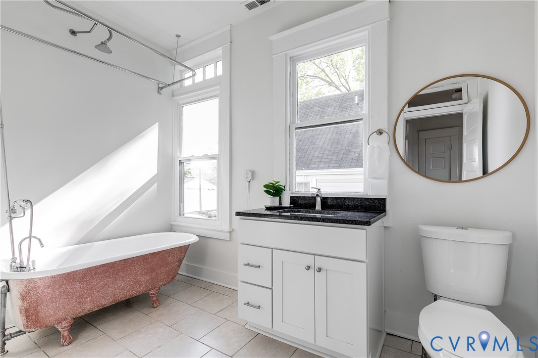 Clawfoot soaking tub with exposed plumbing, white vanity cabinet with stone countertop, large oval mirror, and tile flooring