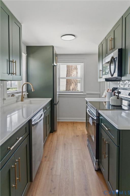 Galley kitchen featuring green cabinetry, gold-tone hardware, stainless steel appliances, light countertops, and wood-finish flooring