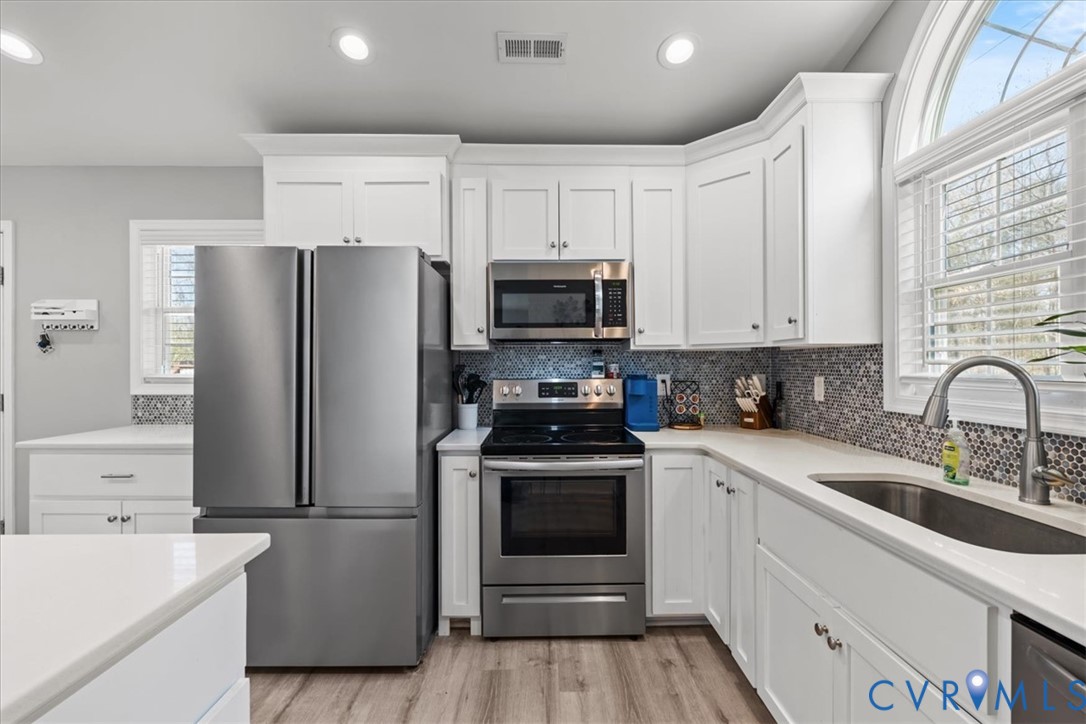 Kitchen featuring white shaker cabinetry, stainless steel appliances, mosaic tile backsplash, solid-surface countertops, and an arched window