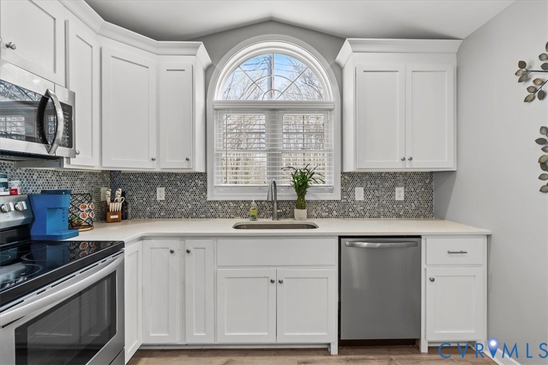 Kitchen featuring white shaker cabinetry, arched window with muntin detailing, stainless steel appliances, apron-front sink, and circular mosaic tile backsplash