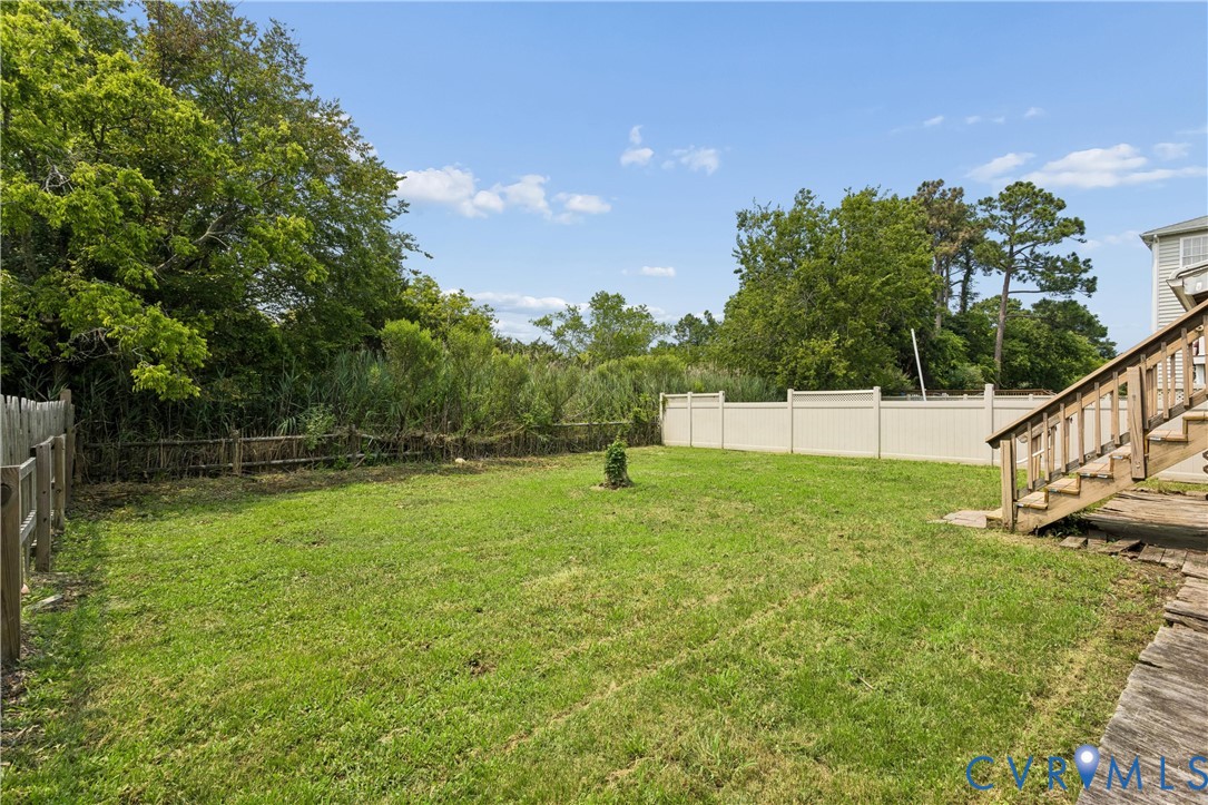 Expansive backyard featuring a manicured lawn, perimeter vinyl fencing, mature trees, and wooden exterior staircase