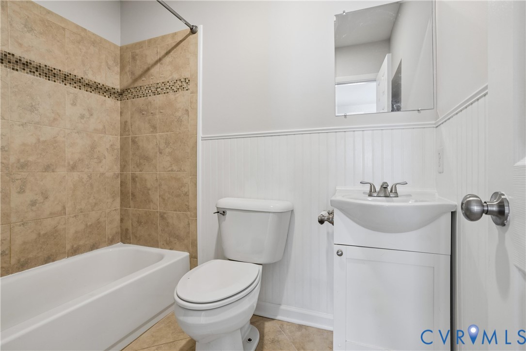 Bathroom featuring a vanity with a pedestal-style sink, white beadboard wainscoting, and a tub enclosure with tan tile and a decorative mosaic accent band