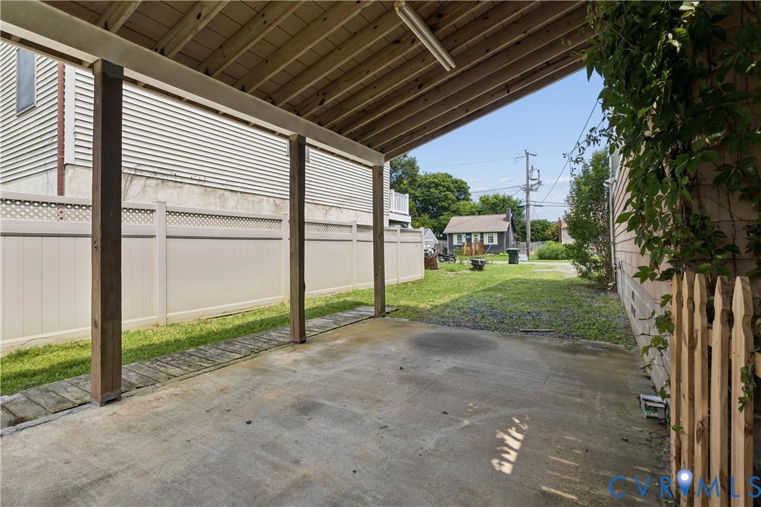 Covered concrete carport featuring exposed wood rafters, vinyl perimeter fencing, and wood-slat partial screen