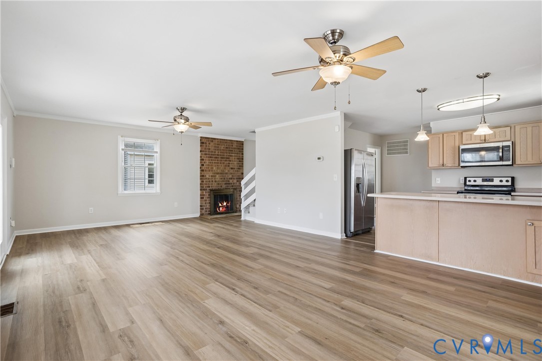 Open-concept living space featuring wood-finish flooring, a full-height brick fireplace, a peninsula kitchen with light-tone cabinetry, and dual ceiling fans Open-concept living space featuring wood-finish flooring, a full-height brick fireplace, a peninsula kitchen with light-tone cabinetry, and dual ceiling fans