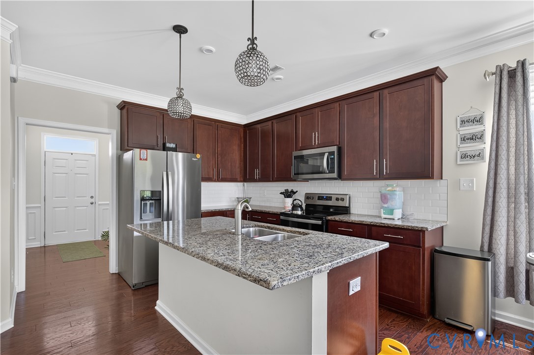 Kitchen featuring wood-finish cabinetry, stone countertops, a center island with a dual-basin sink, subway tile backsplash, and decorative pendant lighting