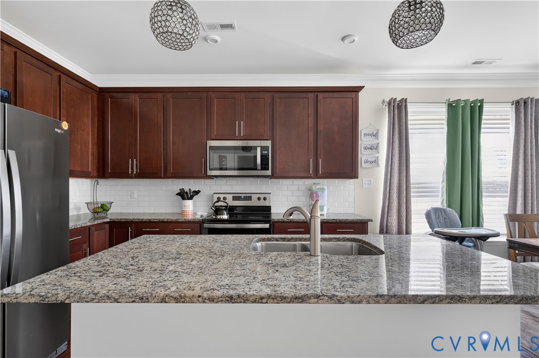 Kitchen featuring dark wood cabinetry, a central island with an integrated stainless steel double-basin sink, stone countertops, a subway tile backsplash, and two spherical pendant lights