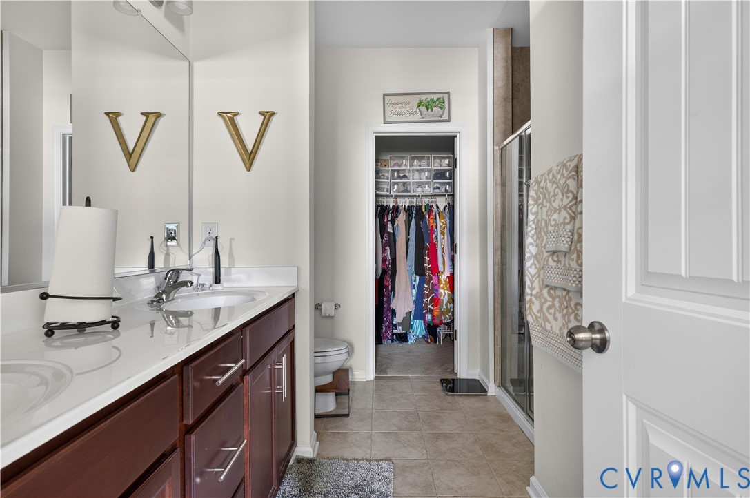 Double vanity featuring a light-toned countertop with integrated dual sinks, dark wood-finish cabinetry, full-width mirror, glass-enclosed shower, and tile flooring