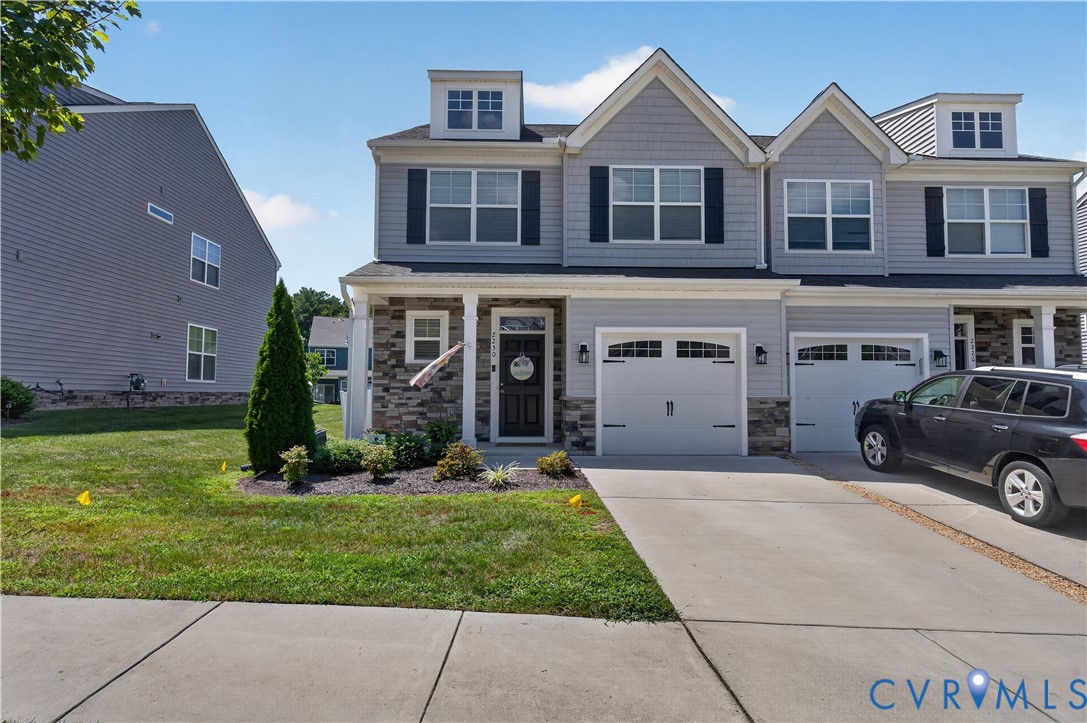 Multi-story townhome exterior featuring gray horizontal siding, stone veneer accents, double gables, dual carriage-style garage doors, and front dormer windows
