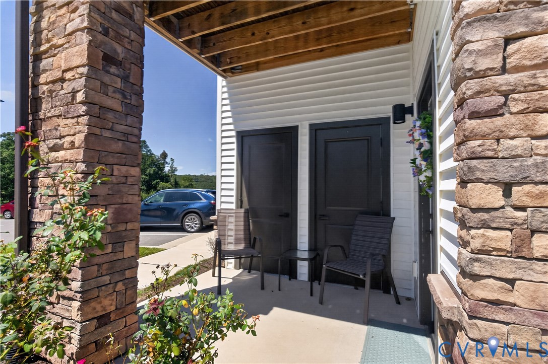 Covered concrete porch featuring stacked stone columns, horizontal siding, and two dark-finish entry doors with wall-mounted lighting