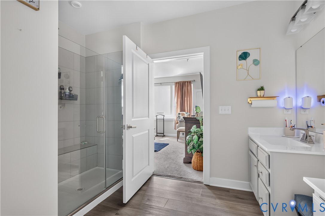 Bathroom featuring a glass-enclosed walk-in shower with tiled surround and integrated bench, wood-finish flooring, vanity with solid-surface countertop, and multi-bulb vanity lighting