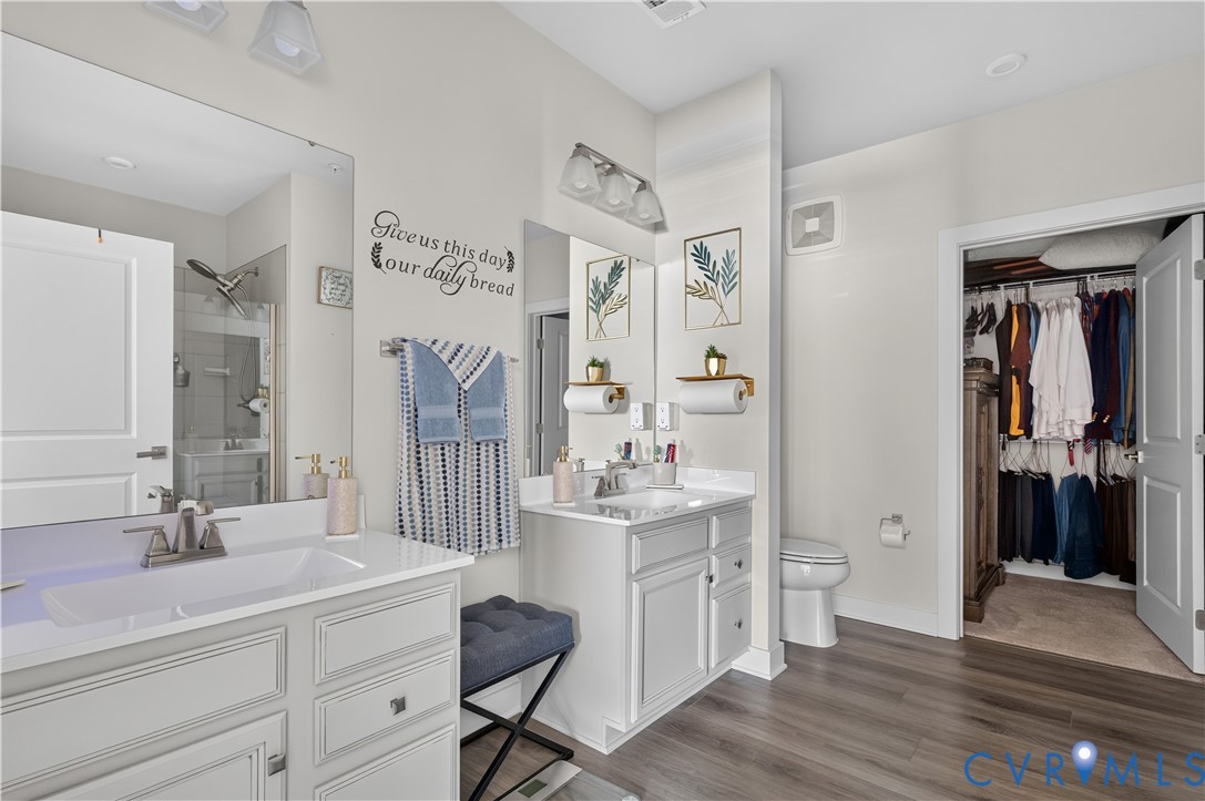 Dual-sink vanity with white cabinetry and solid-surface countertops, full-width vanity mirrors, wood-finish flooring, and walk-in closet with built-in shelving