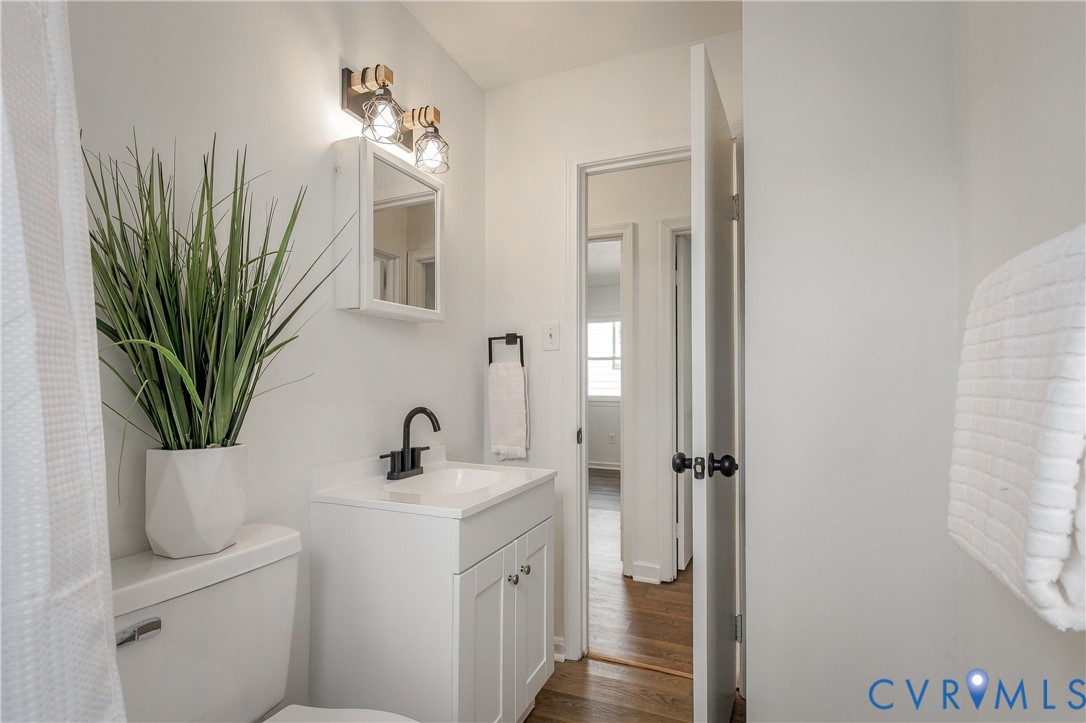 White vanity with a rectangular sink, matte black faucet, wall-mounted medicine cabinet, dual-bulb industrial-style light fixture, and wood-finish flooring