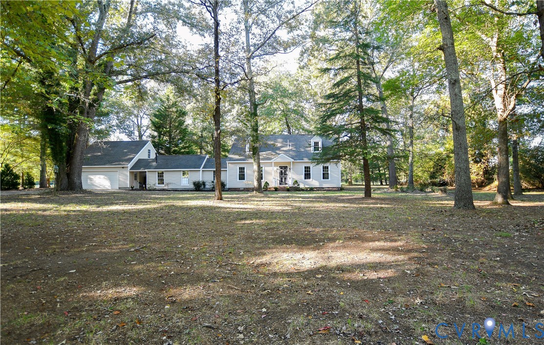 Exterior of a light-colored residence featuring a gabled roof, an attached garage, and mature trees situated on a spacious lot