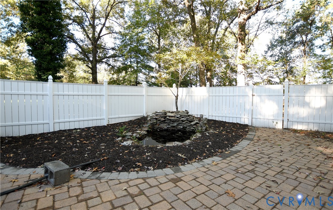 Backyard featuring a white vinyl privacy fence, stone water feature, paved patio area with a circular border, and mature trees