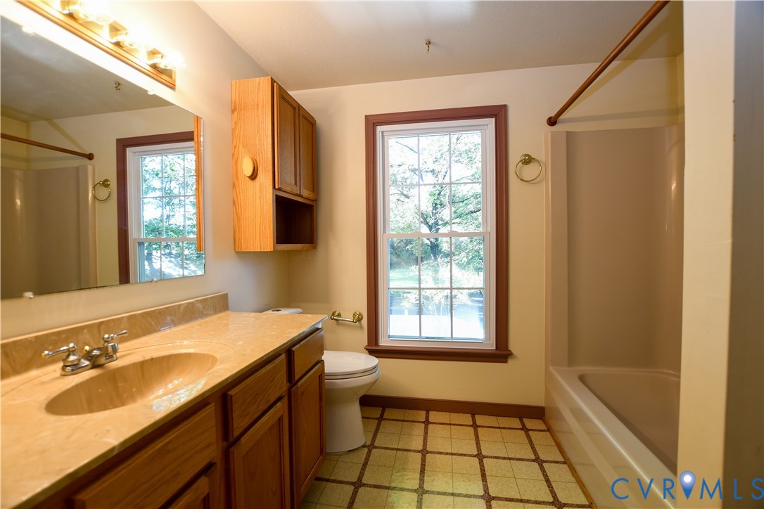 Bathroom featuring a vanity with a solid-surface countertop, integrated sink, wooden cabinetry, wall-mounted mirror with integrated lighting, and a window with a paneled trim