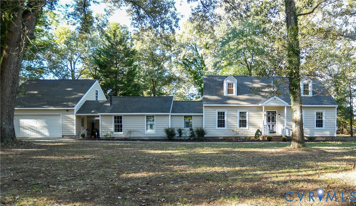 Exterior featuring light-colored horizontal siding, dual front-facing dormer windows, an attached two-car garage, and mature trees across the property grounds Exterior featuring light-colored horizontal siding, dual front-facing dormer windows, an attached two-car garage, and mature trees across the property grounds
