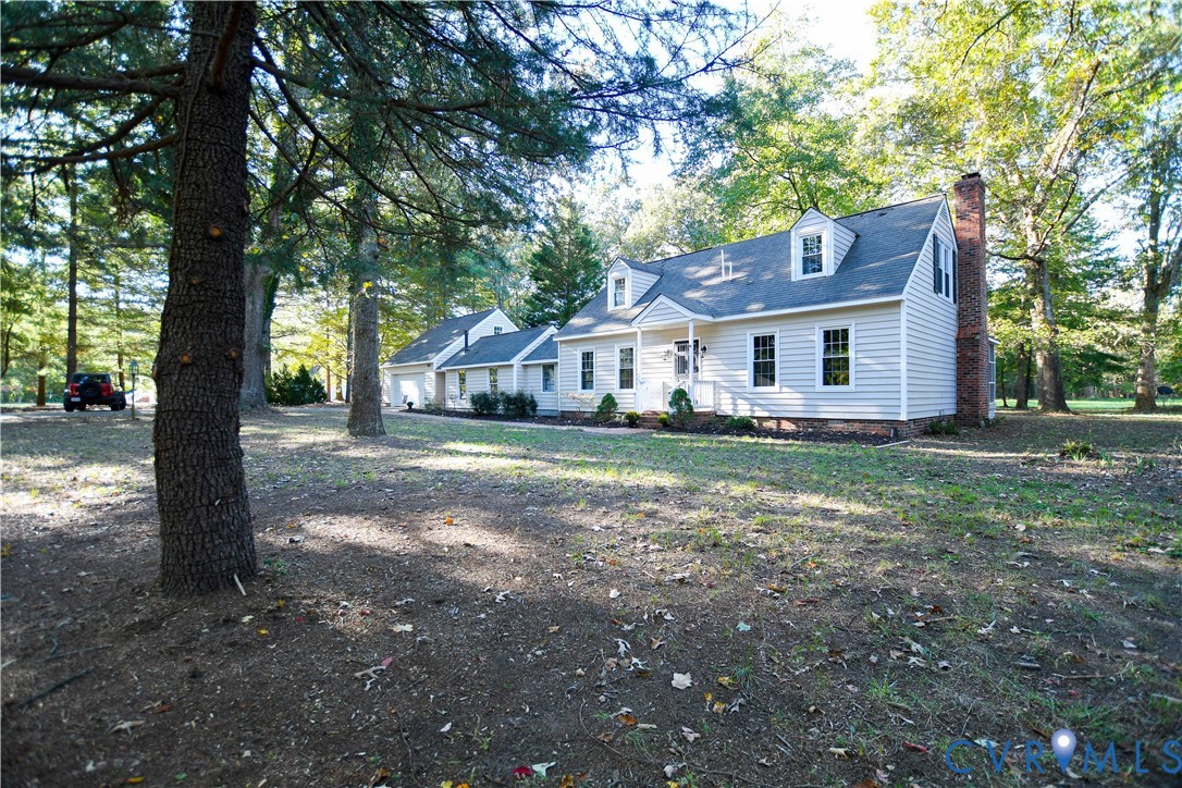 Cape Cod style residence featuring light-colored horizontal siding, dual dormer windows, an exterior brick chimney, and an attached multi-car garage on a wooded lot
