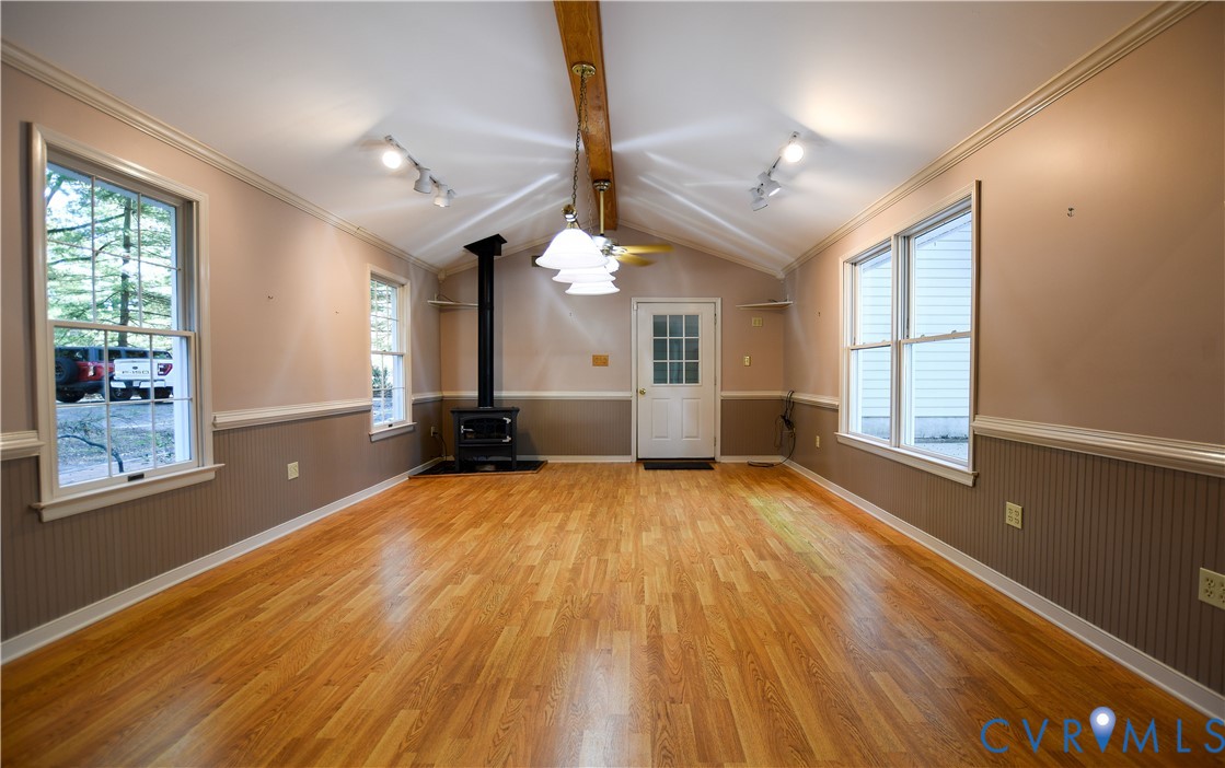 Interior space featuring wood-finish flooring, vaulted ceilings with an exposed wooden beam, beadboard wainscoting, a free-standing metal wood-burning stove, and multiple large windows
