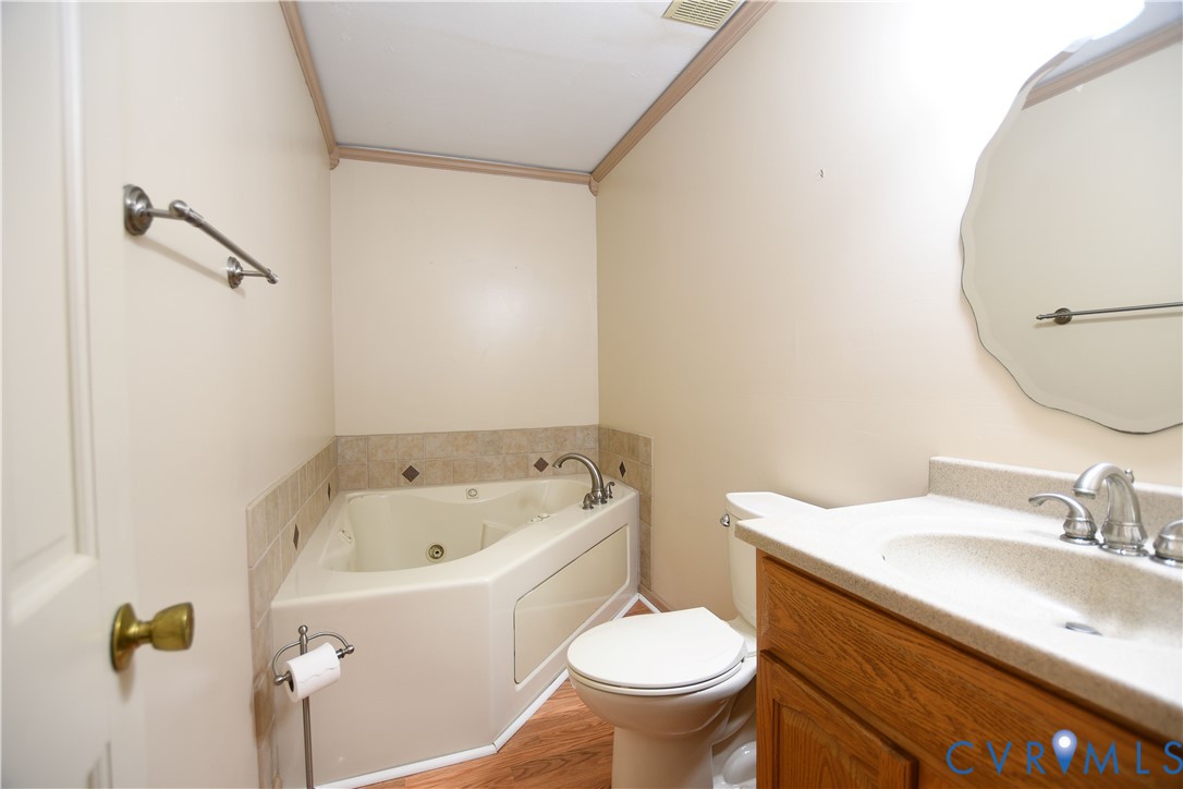 Corner soaking tub with tile surround, wood-finish cabinetry, integrated vanity countertop with sink, and wall-mounted vanity mirror
